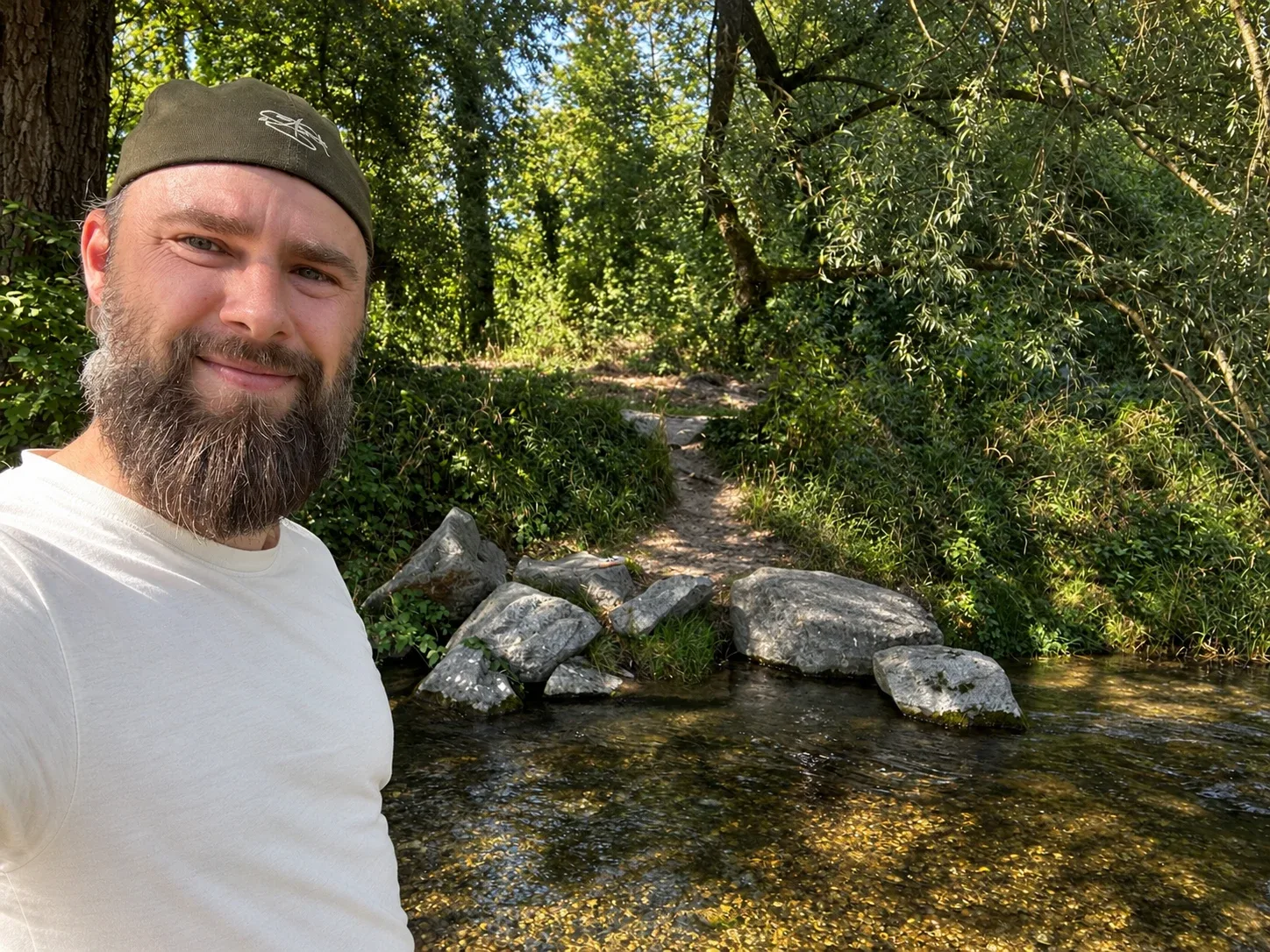 Lasse mit Mütze und langem Bart an einem Bach im Wald.